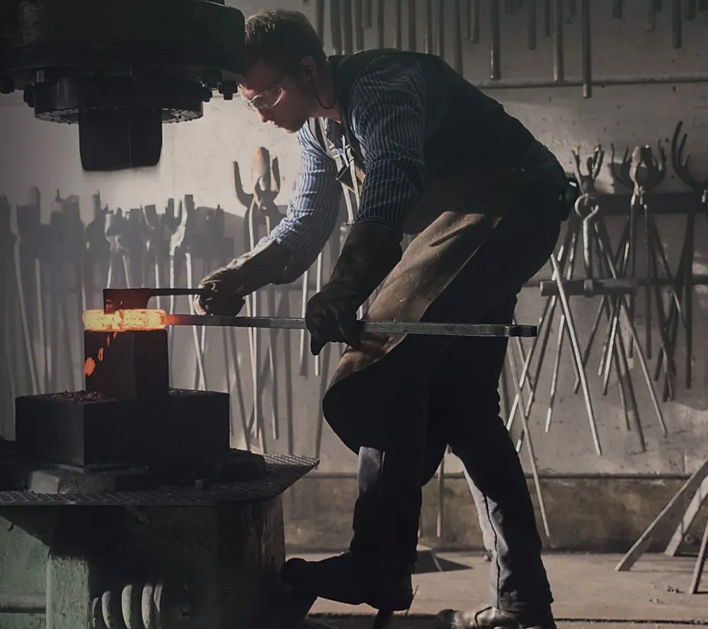 A smith holding a glowing billet under a power hammer